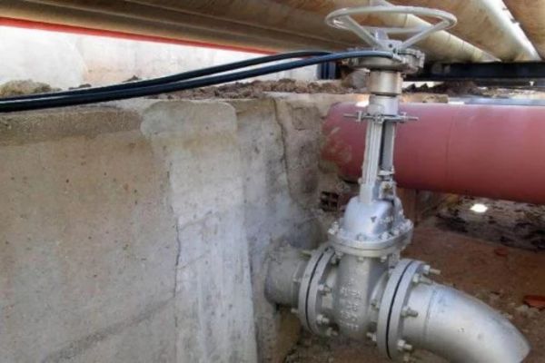 Valvola industriale con volantino e cavo flessibile collegato, montata su una parete in cemento sotto una tubazione.